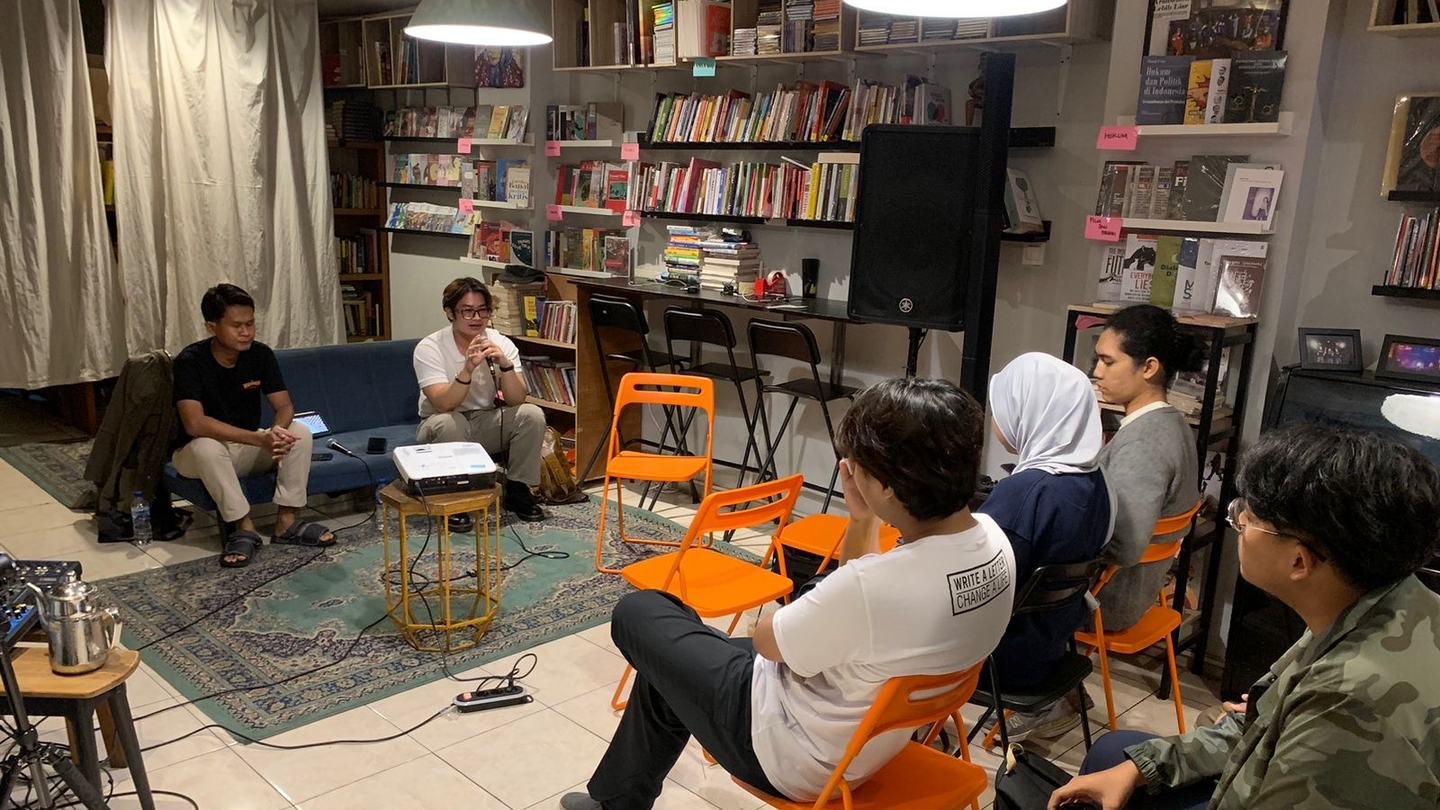 Adha Gozali leading a small discussion in a bookshop, the audience on orange folding chairs.