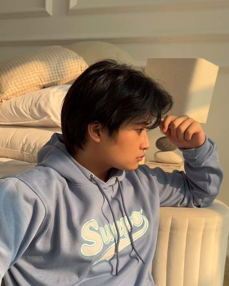 Adha Gozali modelling a light-blue Swepo hoodie, seated against a bed.